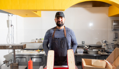 Hands wearing black gloves fold a flatbread filled with sliced turkey, melted cheese, and leafy greens on a checkered paper-lined surface.