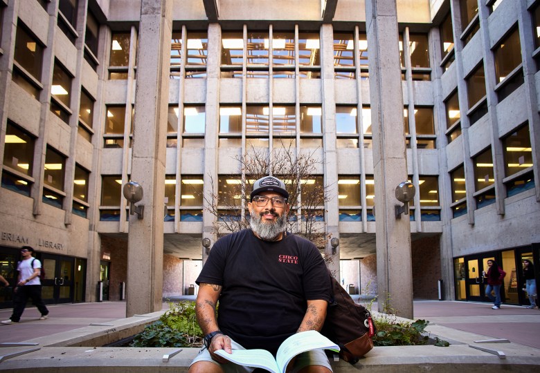 A bearded person wearing a black “Chico State” T-shirt and cap sits on a low concrete planter holding papers, posed in a courtyard framed by tall concrete columns and rows of windows, with a library entrance visible behind them and a few people walking through the background.