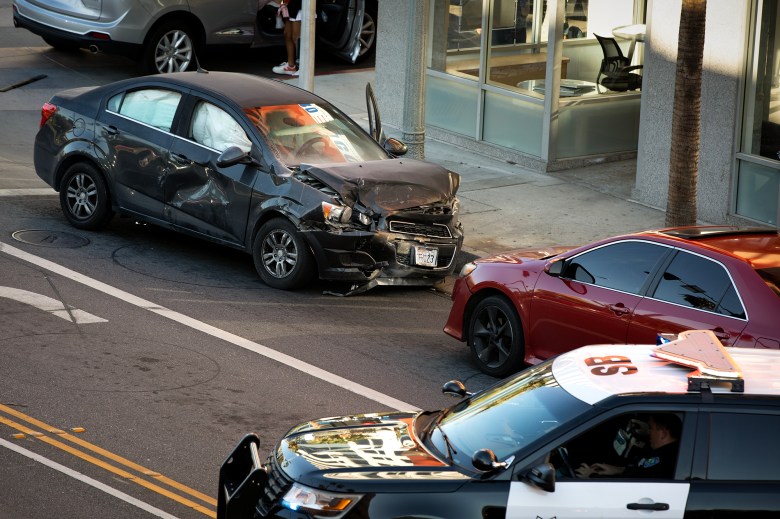 A damaged black sedan with crumpled front and side panels sits in the street after a collision, positioned near a red car while a police vehicle is parked nearby.