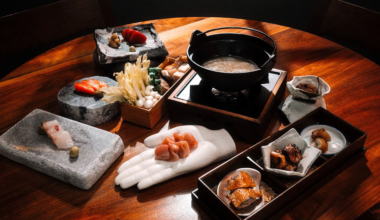 Assorted raw seafood and mushrooms are presented on stone slabs, a ceramic hand, and trays next to a pot of broth on a wooden table.