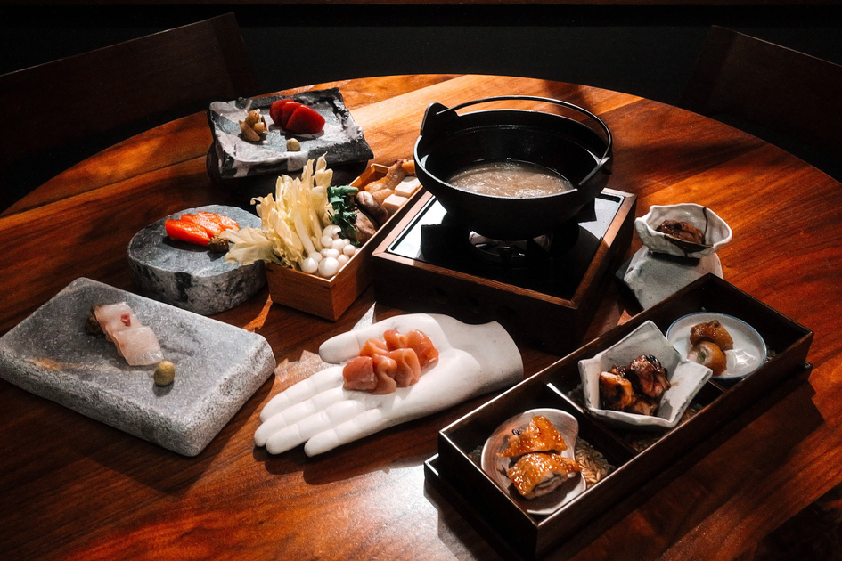 Assorted raw seafood and mushrooms are presented on stone slabs, a ceramic hand, and trays next to a pot of broth on a wooden table.