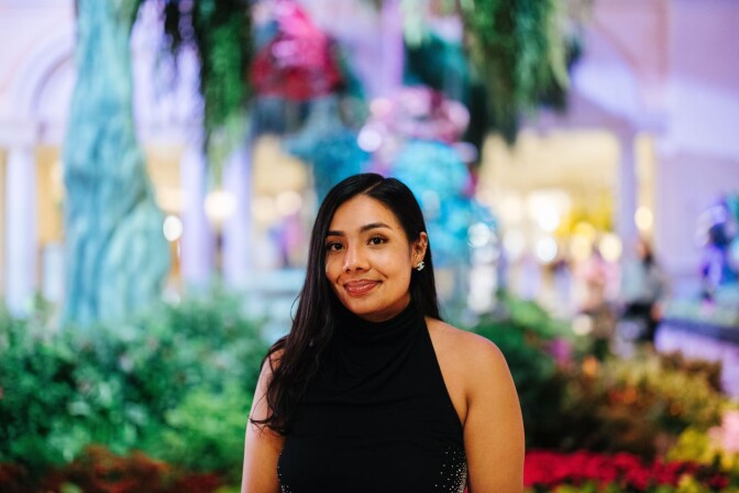 A portrait of a woman with a medium light skin tone wearing a black shoulderless top. Her black hair is long and she's smiling at the camera. The background is blurry, but it's a colorful area.
