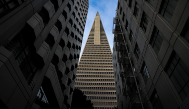 A tall, triangular-shaped skyscraper is framed by two adjacent buildings with multiple windows, with a partly cloudy blue sky visible above.