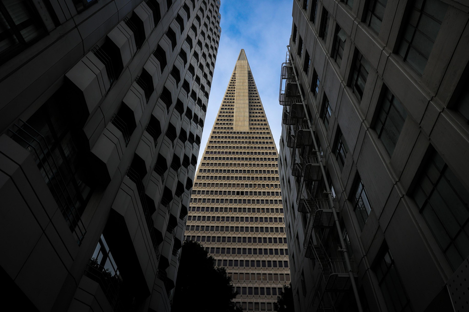 A tall, triangular-shaped skyscraper is framed by two adjacent buildings with multiple windows, with a partly cloudy blue sky visible above.
