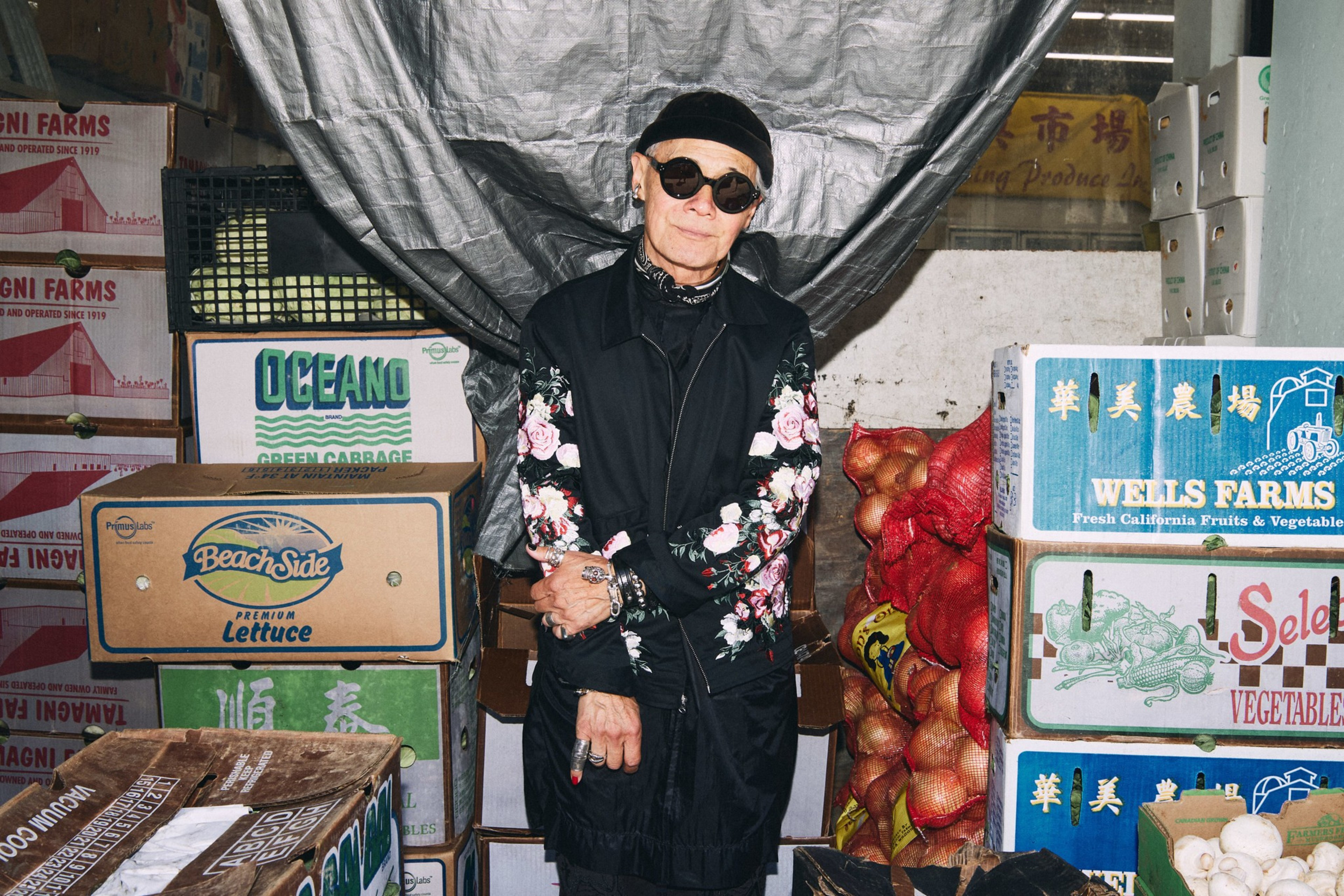 A person in a floral jacket and sunglasses stands among stacked cardboard boxes of produce and mesh bags of onions in a market setting.