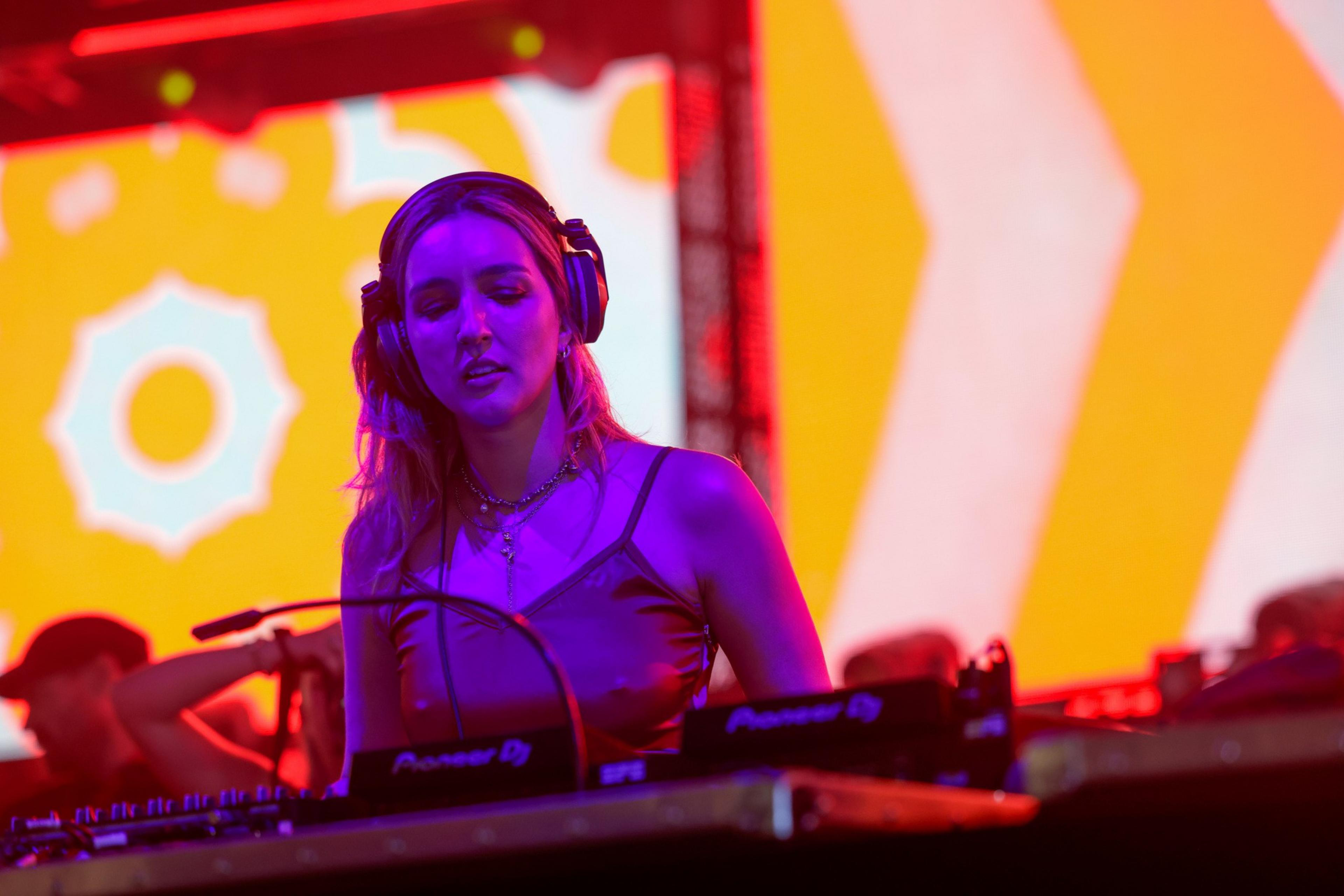 A female DJ wearing headphones mixes music on Pioneer equipment under purple and red stage lights with a bright yellow and white patterned background.