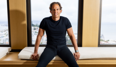 A man with short brown hair wears a black t-shirt and pants, sitting on a cushioned bench in front of large windows showing a cloudy cityscape.
