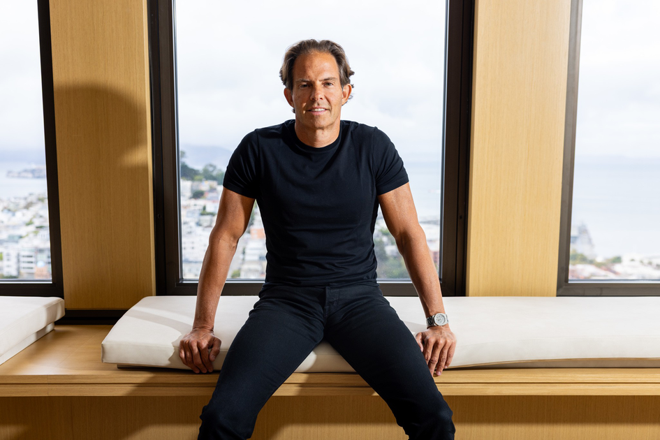 A man with short brown hair wears a black t-shirt and pants, sitting on a cushioned bench in front of large windows showing a cloudy cityscape.