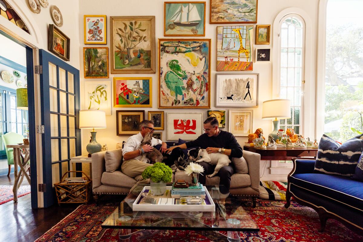 Two men sit on a couch with their three dogs in a living room with high walls covered in paintaings to the ceiling.