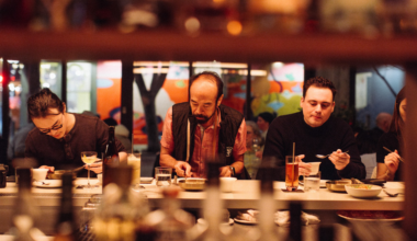 A smiling man with a beard is sitting at a table, holding a camera and looking at its screen, with dishes and a cup in front of him.