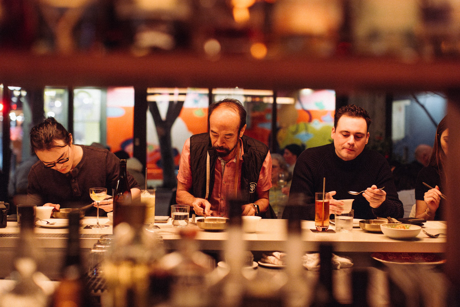 A smiling man with a beard is sitting at a table, holding a camera and looking at its screen, with dishes and a cup in front of him.