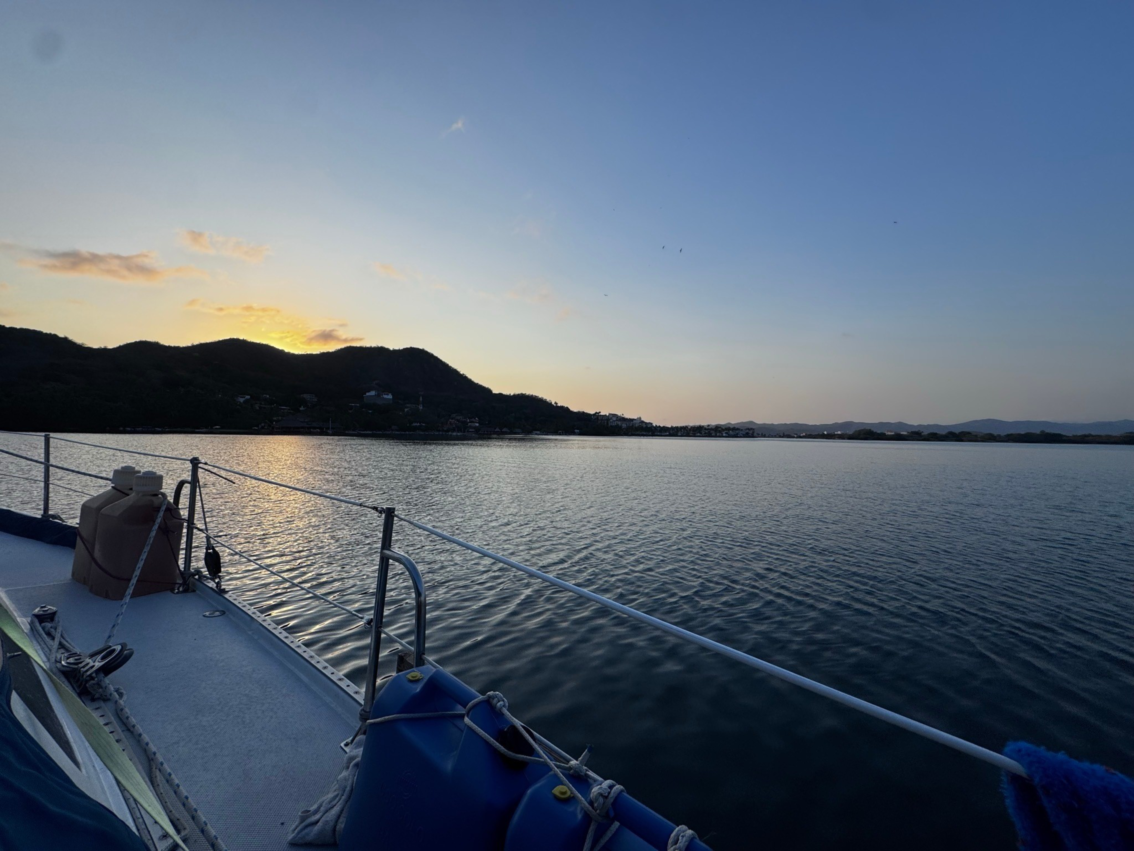 A calm body of water stretches toward silhouetted hills under a clear sky at sunset, viewed from the deck of a boat with railings and equipment visible.