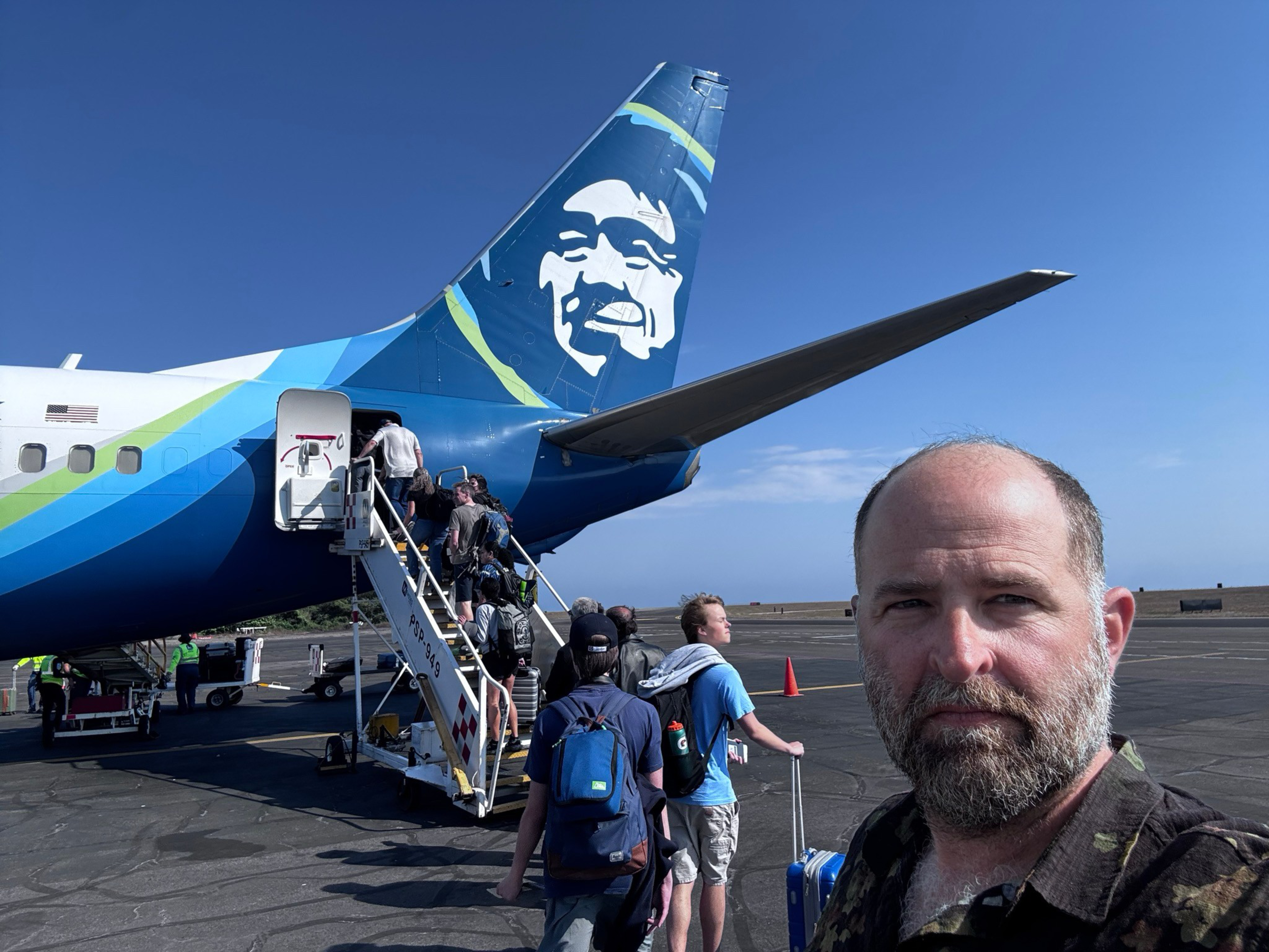A man with a beard takes a selfie while passengers board an Alaska Airlines plane outdoors on a sunny day.