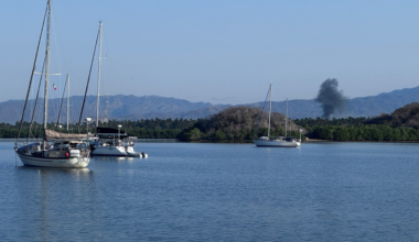 Three sailboats float on calm water near a green, tree-lined shore with mountains in the distance and a plume of black smoke rising against a clear blue sky.