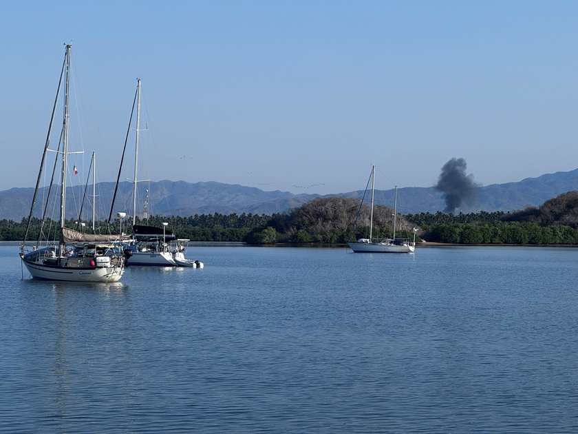 Three sailboats float on calm water near a green, tree-lined shore with mountains in the distance and a plume of black smoke rising against a clear blue sky.