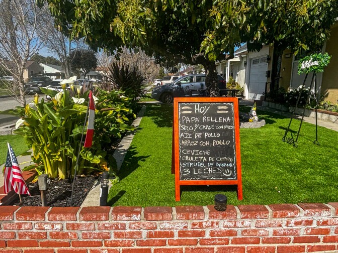 A handwritten chalkboard sign on a front lawn in Lakewood lists the day's specials including Papa Rellena, Seco de Carne con Frijoles, Aji de Pollo, Ceviche, and Tres Leches. A Peruvian flag and American flag are planted in a flower bed nearby