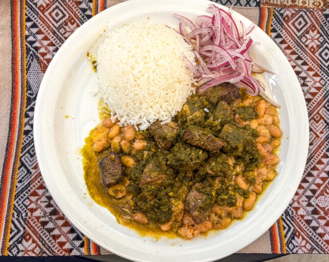 A white plate of Seco de Res con Frijoles featuring tender beef chunks in a deep green cilantro sauce surrounded by canary beans, a mound of white rice, and pickled red onions, served on a colorful Peruvian textile.