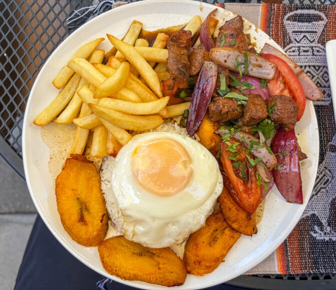 A white bowl of lomo saltado featuring stir fried beef, tomatoes, and red onions served over white rice topped with a fried egg, alongside french fries and fried plantains, on a Peruvian textile.