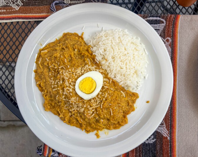 A white plate of Aji de Gallina featuring shredded chicken in a vibrant golden aji amarillo cream sauce, topped with a hard-boiled egg, served alongside white rice on a Peruvian textile.
