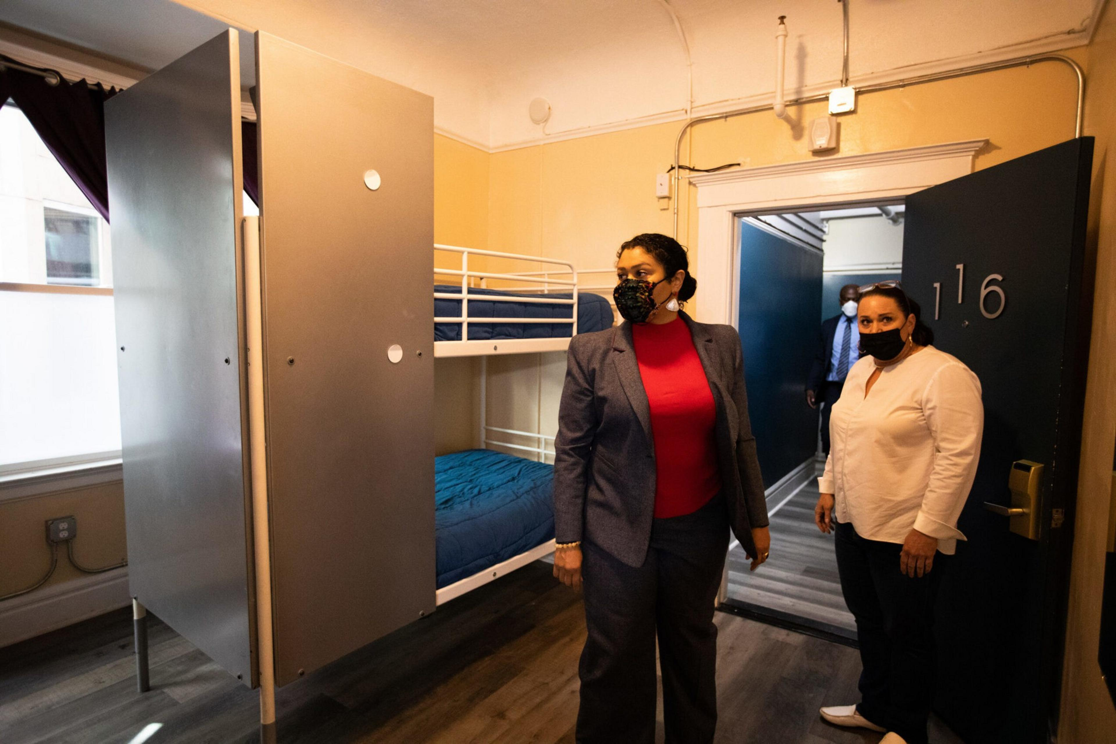 Two women wearing masks stand inside a small room with a bunk bed, open door numbered 116, and a window with a partially drawn curtain.