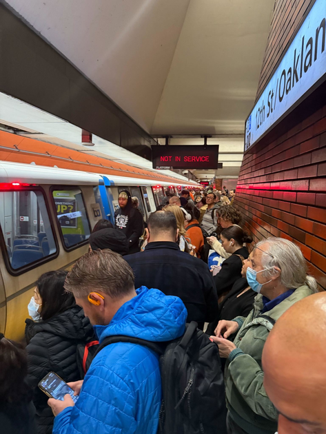 A crowded subway platform at 12th St/Oakland with many people waiting beside a train displaying a "NOT IN SERVICE" sign.
