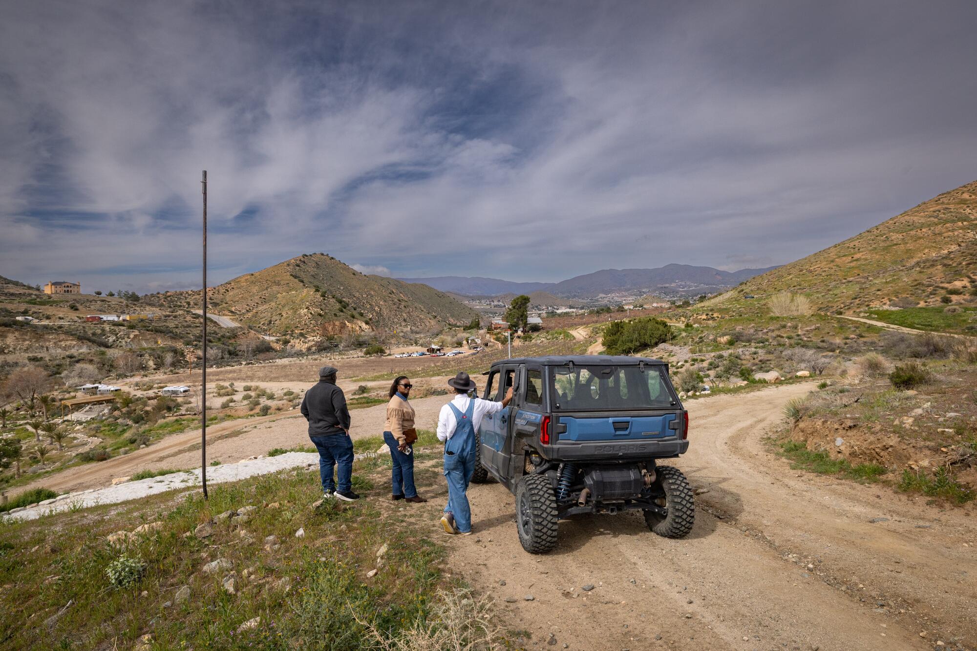 Three people overlook a ranch next to a vehicle.