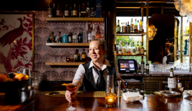 A bartender smiles as she hands over a cocktail inside a warmly lit bar with shelves of liquor and a framed bird artwork.