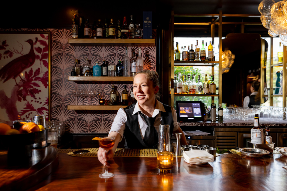 A bartender smiles as she hands over a cocktail inside a warmly lit bar with shelves of liquor and a framed bird artwork.