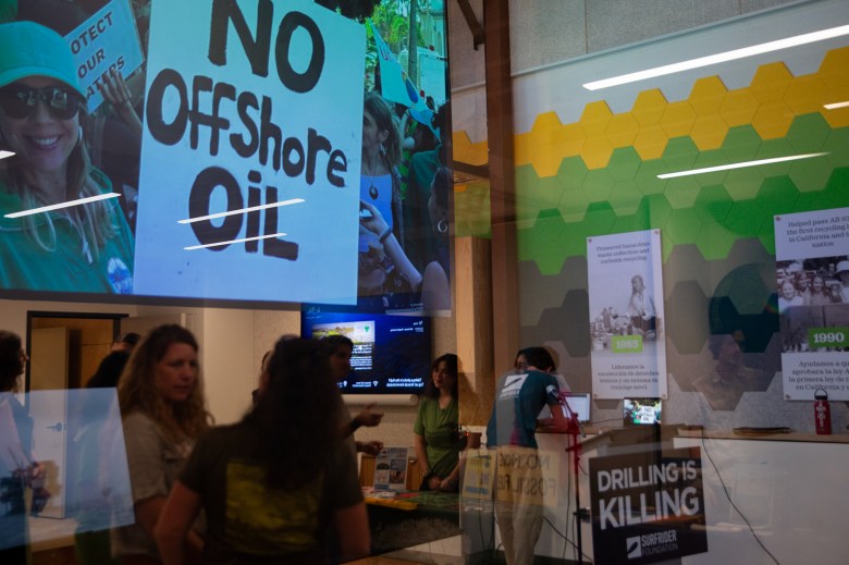 The reflection of people as they stand and mingle during a town hall. A photo of a sign that reads, "No Offshore Oil" is projected onto a screen.
