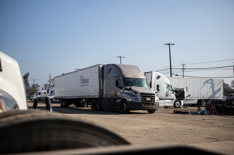 Semi-trucks are parked in a dirt lot, including a gray tractor attached to a refrigerated trailer and a white truck with its hood raised as tools lie on the ground nearby under a clear sky.
