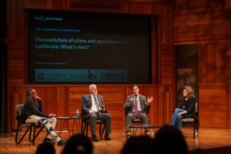 Four people face an audience as they sit on chairs set up on a stage inside an auditorium with mahogany walls. A projector set up behind the four speakers displays a graphic titled "The Evolution of Crime and Punishment in California: What's Next?."