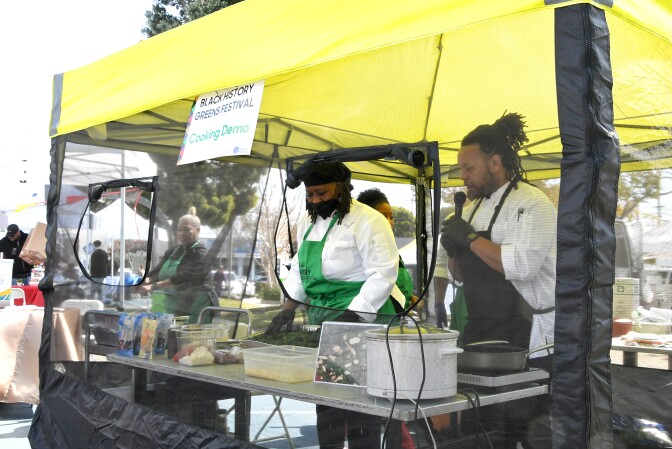 Two Black men cook behind a clear plastic tarp. One also holds a microphone. 