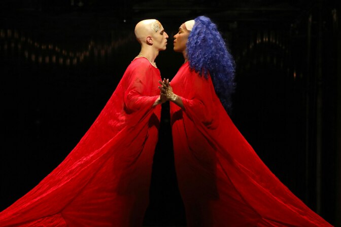 A man with medium skin tone and a shaved head has his face inches from a Black woman with purple hair. They're both wearing long red robes. 