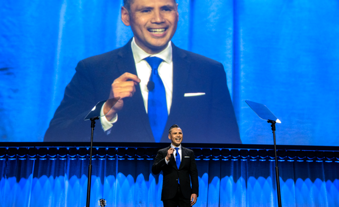 A man with medium skin tone, wearing a black suit and blue tie, speaks on a stage with a large monitor showing him in the backgorund.