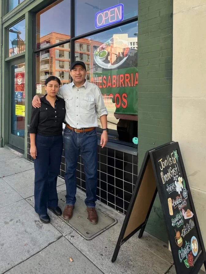 A man and a woman, both with medium skin tone, pose for a photo outside a restaurant next to a chalkboard with writing and illustrations on it.