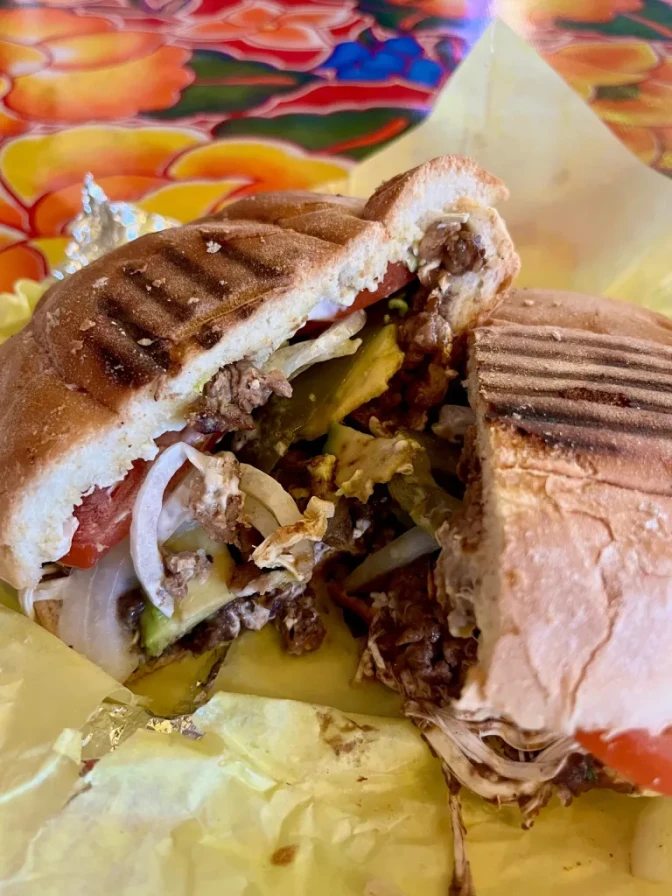 A close up of a torta cut in half with vegetables and meat spilling outside of it on yellow deli paper on a floral painted table.