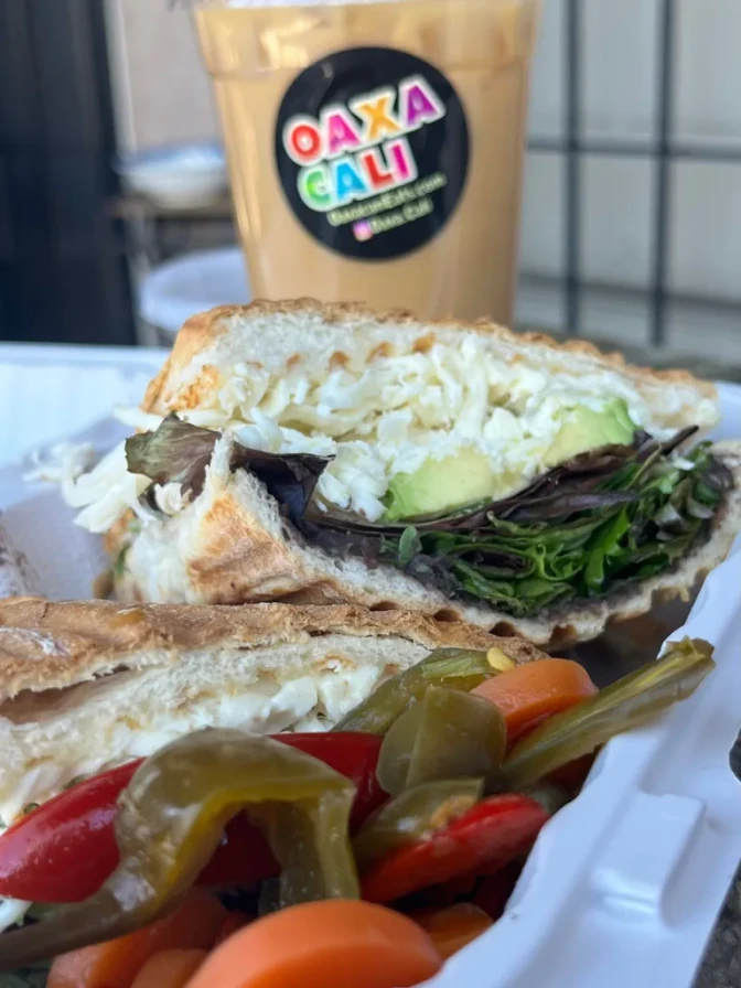 A torta with a lot of vegetables and cheese, next to an iced coffee drink with a sticker that reads "OaxacaCali."