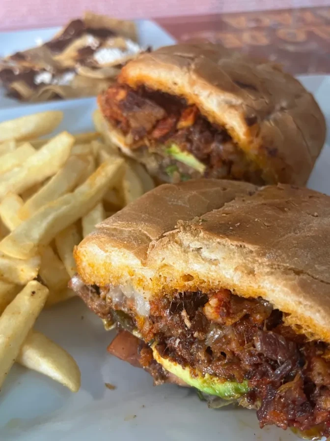 A close up of a torta with meat, beans, and avocado, cut in half, next to a side of fries.