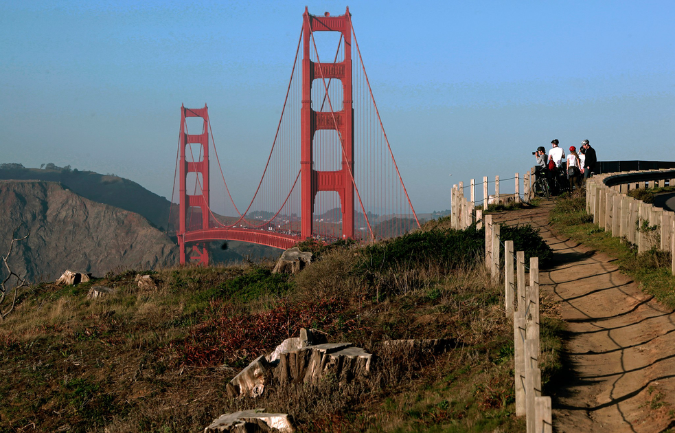 A group of people stands on a fenced dirt path overlooking the iconic red Golden Gate Bridge with hills and a clear blue sky in the background.