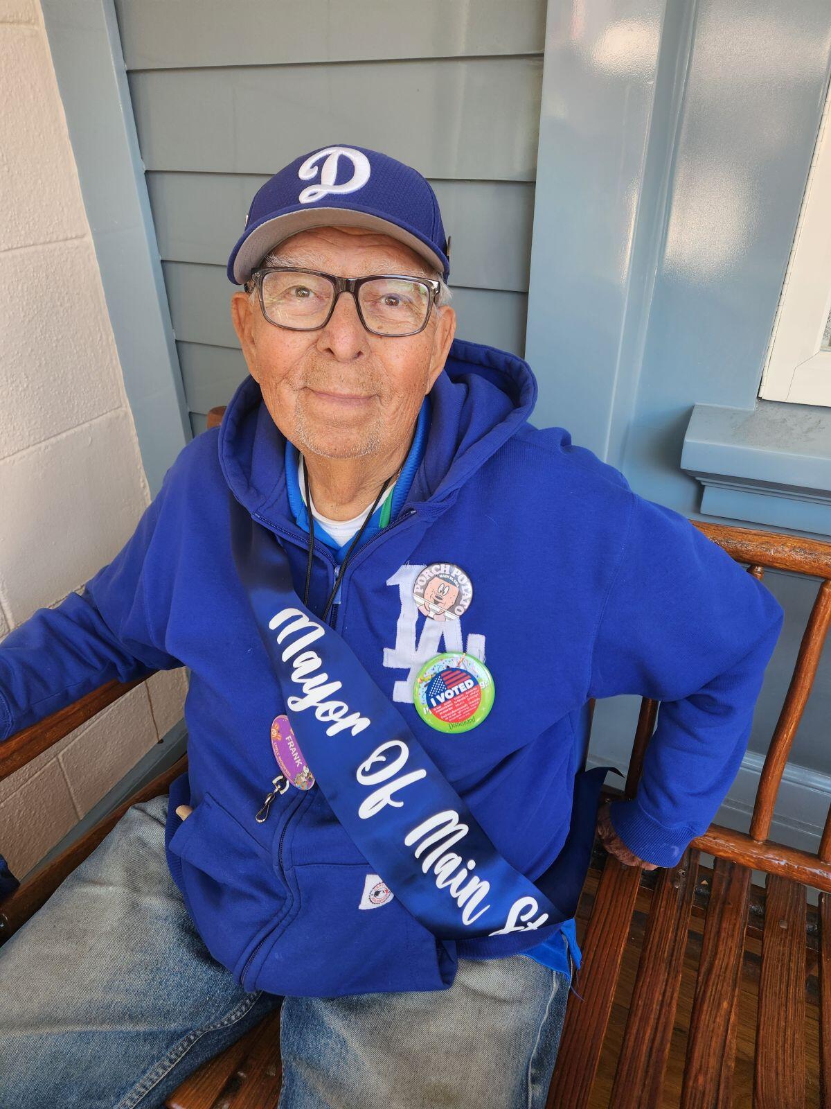 A man in glasses and a blue cap with a cursive "D" sits on a bench  
