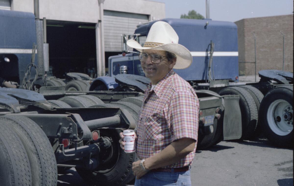 A man in a light colored cowboy hat drinks a canned beverage near a  large truck.