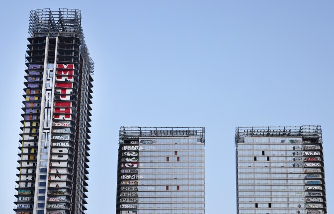Three unfinished skyscrapers with scaffolding and graffiti-covered facades stand against a clear blue sky