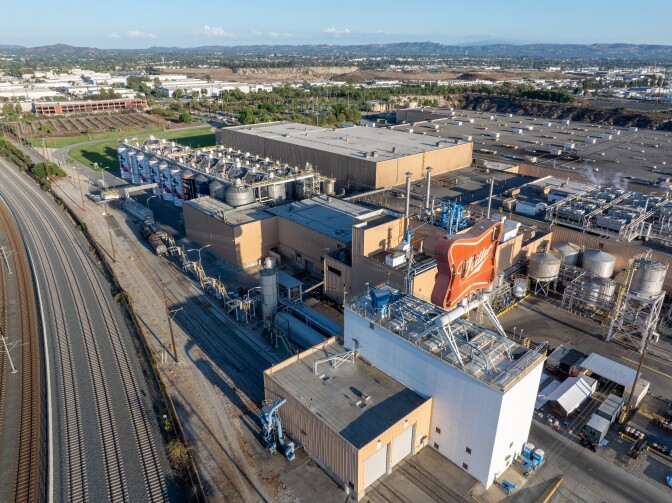 An aerial view of the Irwindale brewery taken in September 2025, after the January winds tore up the sign. The red "Miller" sign is seen above the rest of the brewery. Mountains and the sky can be seen farther in the background. Train tracks can be seen to the left of the brewery.