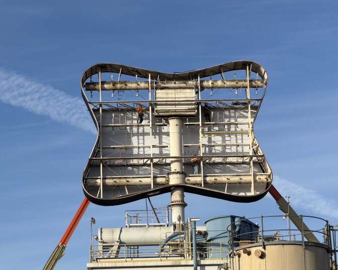 Workers can be seen inside the Irwindale brewery sign as he works on replacing the old "Miller" sign with the new "City Brewing & Beverage Irwindale" sign. Construction cranes are seen on both sides of the sign. There's is a clear blue sky behind the sign.