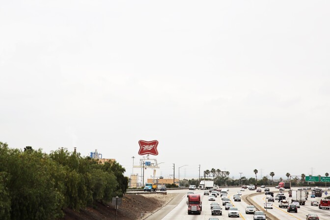 A view of the old red "Miller" sign from the 210 Freeway on a hazy day. Traffic flows in both directions as commuters drive by the brewery. Palm trees can be seen near the horizon.