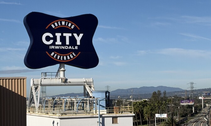 The new Irwindale brewery sign as its seen from the 210 freeway. It is large and blue and says "City Brewing & Beverage Irwindale." Mountains and a blue sky can be seen behind it. Train tracks can be seen to the right of it. The rest of the Irwindale brewery is seen peeking out to the left of the sign.