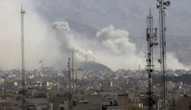 A group of people wearing purple shirts stand on a sidewalk outside a building, holding signs that say "SEIU Unfair Labor Practice On Strike."