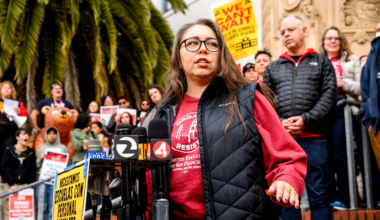 A woman wearing glasses and a red sweatshirt speaks at a protest with microphones nearby and people holding signs behind her.