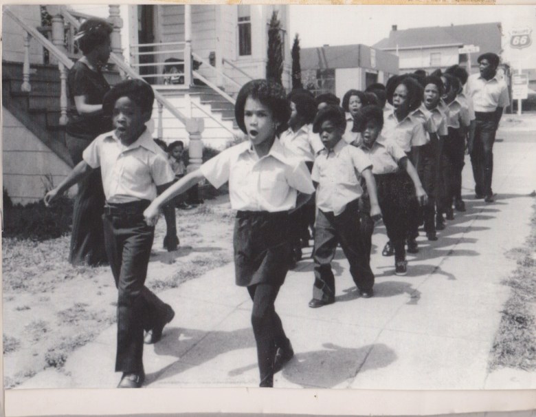 A historic, black and white photograph depicts a line of children in black berets and matching outfits march in formation down a street.