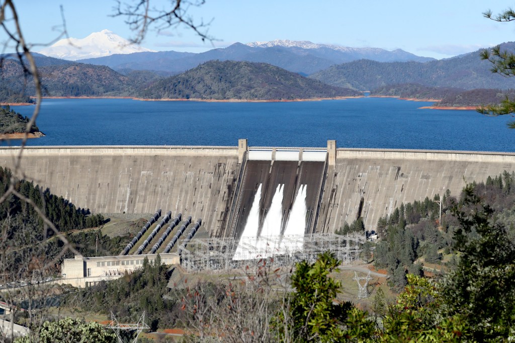 Water flows from five gates on Shasta Dam with a lake behind it and snowy mountains in the distance.
